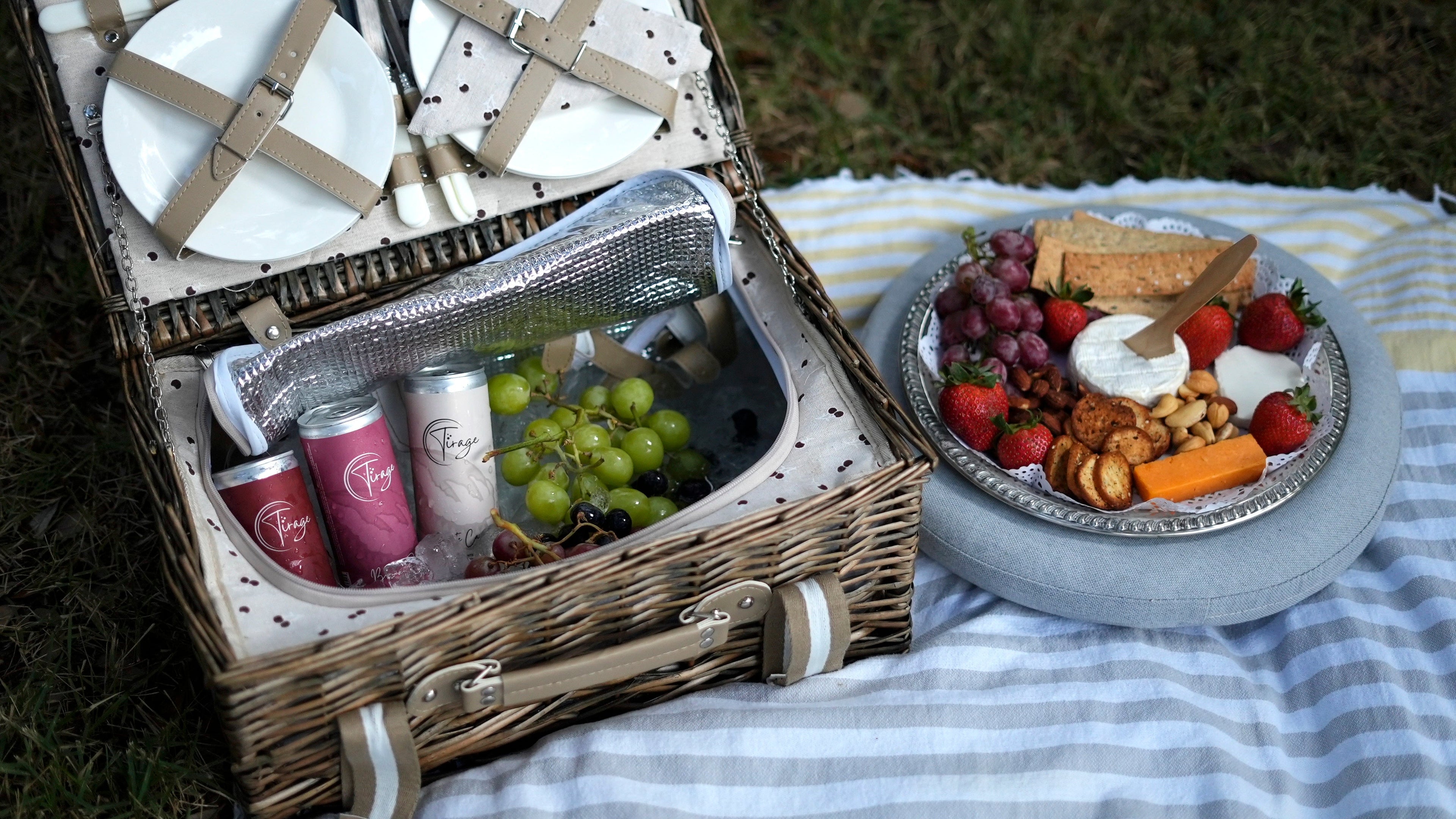 Wicker picnic basket with plates, drinks, grapes and cheese platter on striped blanket outdoors