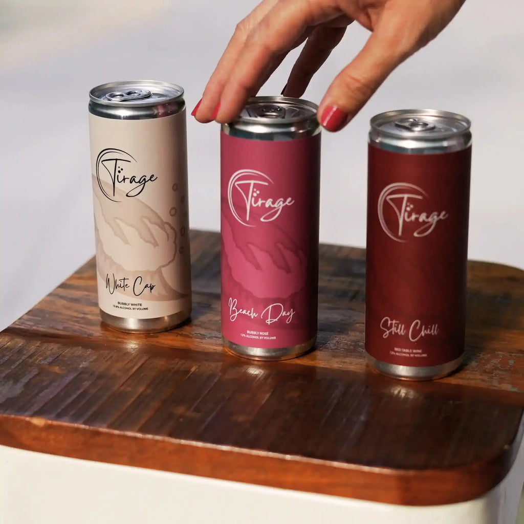 Three Tirage canned drinks on a wooden surface with a hand reaching for one can.