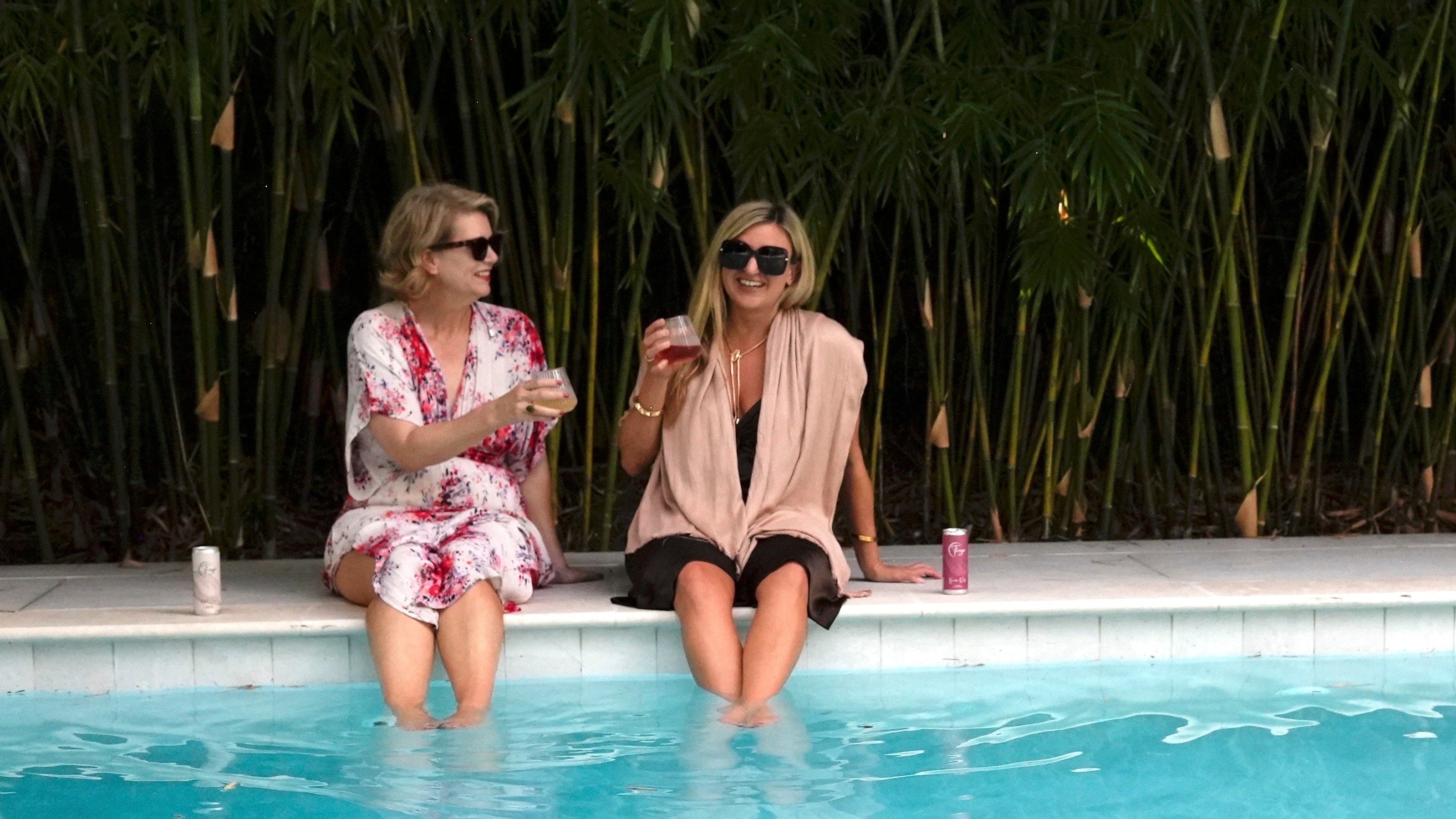 Two women sitting by a pool with drinks, surrounded by greenery.